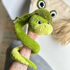 Puede incluir: Juguete de serpiente de crochet verde con cabeza de rana. La rana tiene ojos negros y un sombrero verde con una estrella blanca.