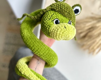 Puede incluir: Juguete de serpiente de crochet verde con cabeza de rana. La rana tiene ojos negros y un sombrero verde con una estrella blanca.