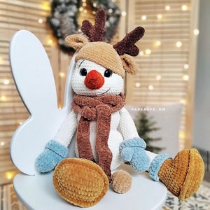 Puede incluir: Un muñeco de nieve de peluche hecho a mano con sombrero marrón, cuernos y bufanda. El muñeco de nieve tiene una nariz de zanahoria, mitones azules y botas marrones. Un pequeño objeto redondo de color marrón amarillento está delante.