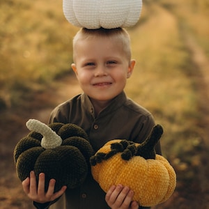 Könnte beinhalten: Ein Kind hält zwei Plüschkürbisse, einen dunkelgrünen und einen gelben, mit grünen Stielen. Ein weißer Kürbis mit braunem Stiel ruht auf dem Kopf des Kindes. Die Kürbisse sind handgefertigt und wahrscheinlich für die Herbstdekoration.