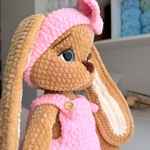 May include: A plush crochet rabbit doll with tan fur, long ears, and a pink headband with a bow. The doll wears pink overalls with a small wooden button on the chest. It features large blue eyes with long black eyelashes and a dark brown nose.