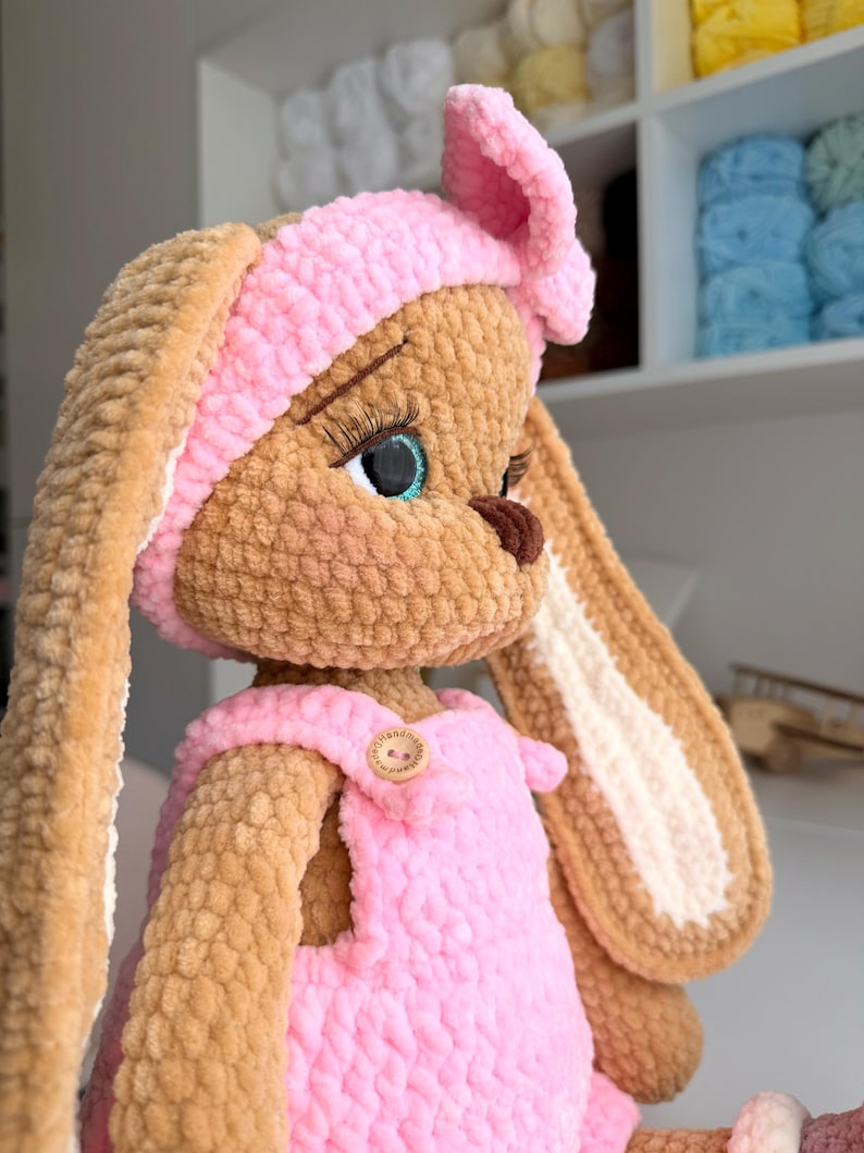 May include: A plush crochet rabbit doll with tan fur, long ears, and a pink headband with a bow. The doll wears pink overalls with a small wooden button on the chest. It features large blue eyes with long black eyelashes and a dark brown nose.