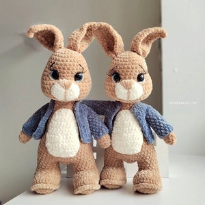 Puede incluir: Dos peluches de conejo marr&oacute;n de crochet con chaquetas azules. Los conejos tienen caras blancas y ojos azules.