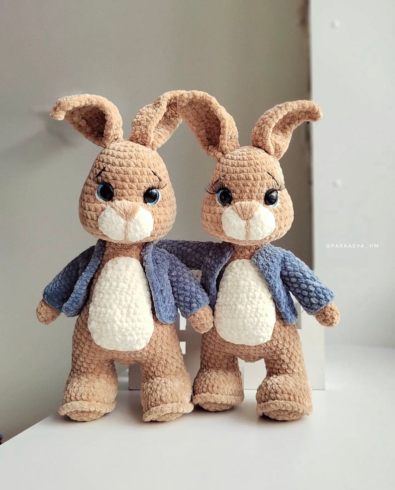 Puede incluir: Dos peluches de conejo marr&oacute;n de crochet con chaquetas azules. Los conejos tienen caras blancas y ojos azules.