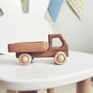 May include: A toy truck crafted from dark brown wood with a visible grain pattern, featuring a flatbed cargo area, a cabin with a cutout window, and four light-colored, natural wood wheels with a circular design on each side of the vehicle.