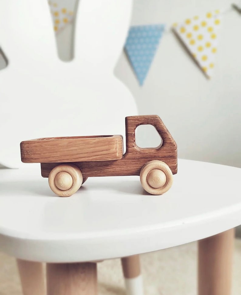 May include: A toy truck crafted from dark brown wood with a visible grain pattern, featuring a flatbed cargo area, a cabin with a cutout window, and four light-colored, natural wood wheels with a circular design on each side of the vehicle.