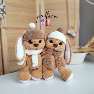 May include: Two crocheted bunny plushies with white fur and brown accents. One bunny is wearing a white hat and a brown scarf, while the other bunny is wearing a brown hat and a white scarf. The bunnies are sitting on a wooden surface.