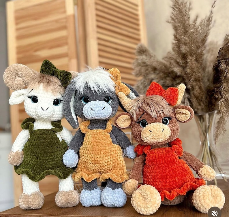 May include: Three crocheted stuffed animals: a white lamb with green overalls, a gray donkey with a yellow dress, and a brown cow with a red dress.