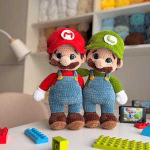 Puede incluir: Dos muñecos de peluche tejidos a mano que representan a Mario y Luigi. Mario lleva una gorra roja con una M, camisa roja, peto azul y zapatos marrones. Luigi lleva una gorra verde con una L, camisa verde, peto azul y zapatos marrones. Ambos tienen guantes blancos.