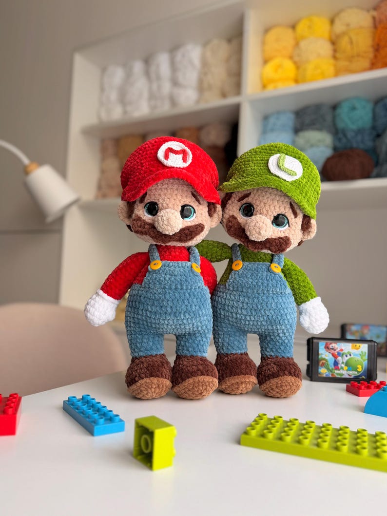 May include: Two handmade crochet plush dolls representing Mario and Luigi. Mario wears a red hat with an M, red shirt, blue overalls, and brown shoes. Luigi wears a green hat with an L, green shirt, blue overalls, and brown shoes. Both have white gloves.