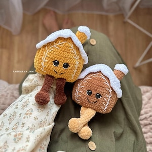 Op de afbeelding: Twee gehaakte gingerbread house speelgoedjes met lachende gezichten. De huizen zijn bruin en geel met witte gehaakte daken. De huizen hebben bruine en beige gehaakte poten.