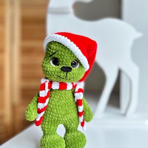 Peut inclure: Une peluche crochet&eacute;e verte &agrave; la texture douce, dot&eacute;e de grands yeux sombres et d'un nez noir brod&eacute;. Le jouet porte un bonnet de P&egrave;re No&euml;l rouge et blanc ainsi qu'une &eacute;charpe assortie &agrave; rayures rouges et blanches autour du cou.
