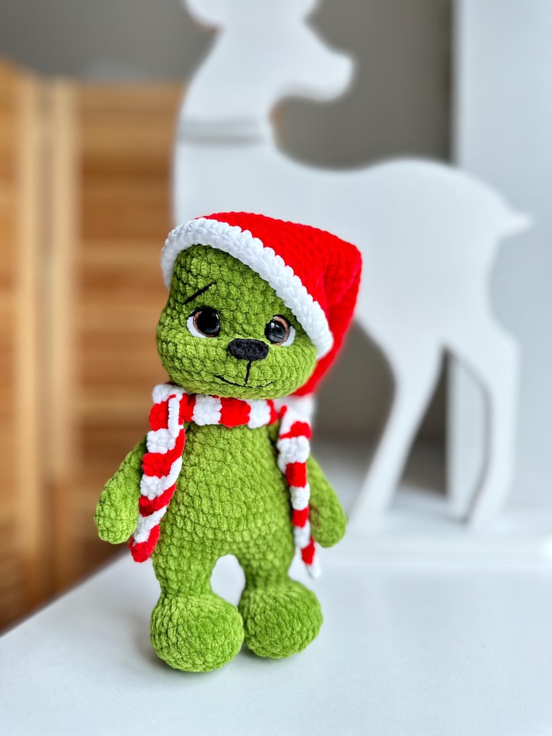 Peut inclure: Une peluche crochet&eacute;e verte &agrave; la texture douce, dot&eacute;e de grands yeux sombres et d'un nez noir brod&eacute;. Le jouet porte un bonnet de P&egrave;re No&euml;l rouge et blanc ainsi qu'une &eacute;charpe assortie &agrave; rayures rouges et blanches autour du cou.