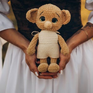 May include: A handmade, tan-colored crochet doll with large ears, a small nose, and green eyes. The doll is wearing a cream-colored outfit. The doll is held in front of a white dress.