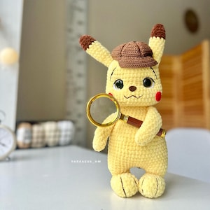 Può includere: Una figura di peluche all'uncinetto gialla che ricorda un detective, indossa un cappello da cacciatore marrone e tiene in mano una lente d'ingrandimento con bordo dorato e manico marrone. La figura ha grandi occhi neri, guance rosse e il testo PARKAEVA_HM.