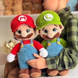 May include: Two handmade crochet plush dolls representing Mario and Luigi. Mario wears a red hat with an M, a red shirt, and blue overalls. Luigi wears a green hat with an L, a green shirt, and blue overalls. Both have brown mustaches and white gloves.