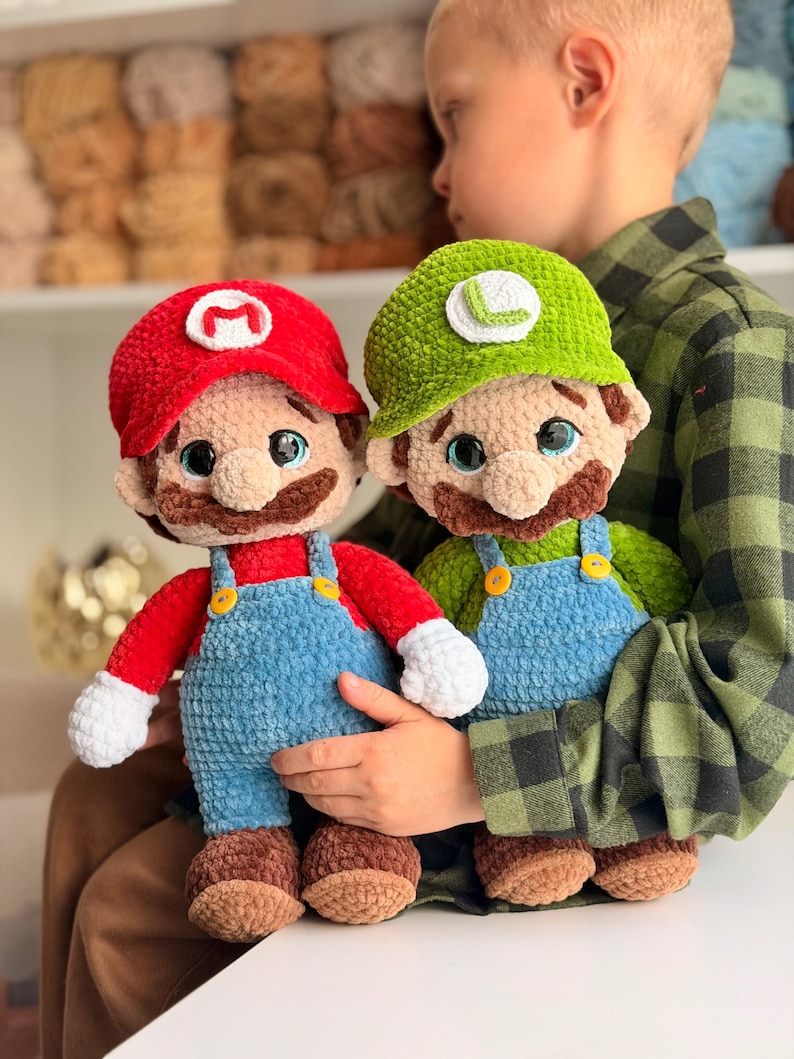 May include: Two handmade crochet plush dolls representing Mario and Luigi. Mario wears a red hat with an M, a red shirt, and blue overalls. Luigi wears a green hat with an L, a green shirt, and blue overalls. Both have brown mustaches and white gloves.
