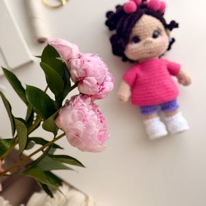 May include: A handmade crochet doll with dark brown hair styled with pink accents, wearing a bright pink short-sleeved shirt, purple shorts, and white shoes, positioned next to a stem of two light pink peony flowers with green leaves.