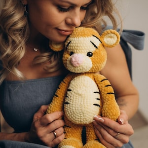 May include: A plush tiger toy crafted from soft, golden-orange yarn with a cream-coloured belly and muzzle. The stuffed animal features black embroidered stripes, a small pink nose, round ears, and dark eyes, showcasing a textured, crocheted finish.