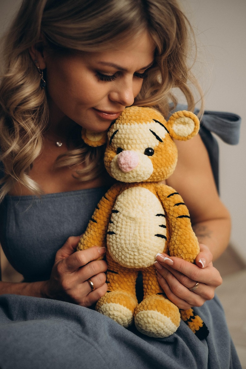 May include: A plush tiger toy crafted from soft, golden-orange yarn with a cream-coloured belly and muzzle. The stuffed animal features black embroidered stripes, a small pink nose, round ears, and dark eyes, showcasing a textured, crocheted finish.