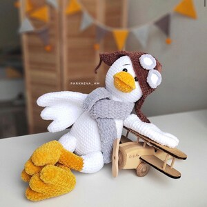May include: A white crocheted bird wearing a brown hat with goggles and a gray scarf, sitting next to a wooden toy airplane.