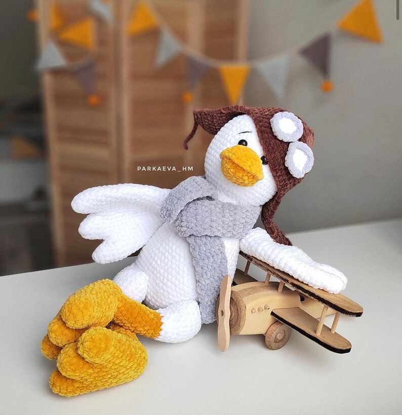 May include: A white crocheted bird wearing a brown hat with goggles and a gray scarf, sitting next to a wooden toy airplane.