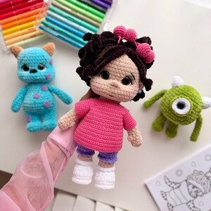 May include: A handmade crochet doll with dark brown curly hair, pink heart hair accessories, a bright pink t-shirt, purple shorts, and white shoes. The doll is accompanied by a blue monster crochet toy and a green one-eyed monster crochet toy.