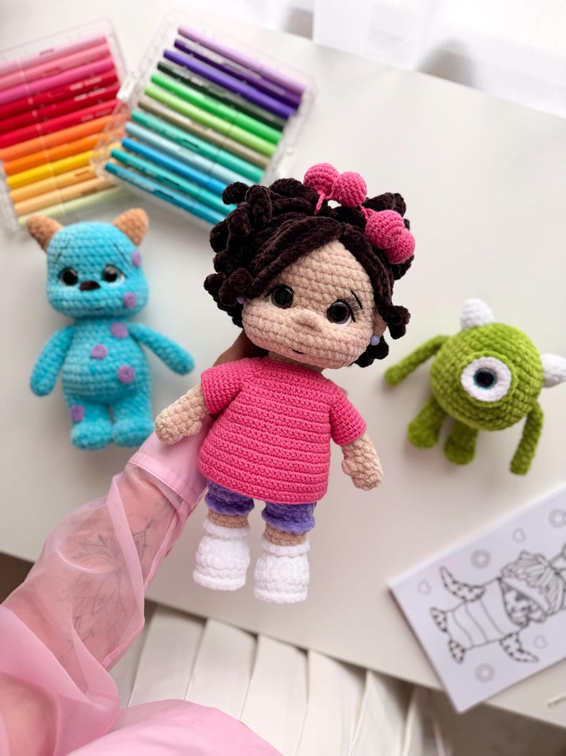 May include: A handmade crochet doll with dark brown curly hair, pink heart hair accessories, a bright pink t-shirt, purple shorts, and white shoes. The doll is accompanied by a blue monster crochet toy and a green one-eyed monster crochet toy.