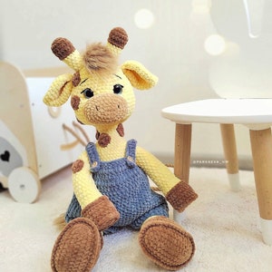 May include: A crocheted giraffe toy wearing a blue denim overall. The giraffe has brown spots and a yellow body. The toy is sitting on a white rug.