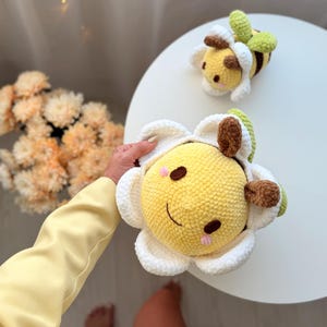 K&ouml;nnte beinhalten: Zwei handgefertigte H&auml;keltiere: eine gelbe Biene und eine gelbe Blume mit einem l&auml;chelnden Gesicht. Die Blume hat wei&szlig;e Bl&uuml;tenbl&auml;tter und braune Akzente. Die Biene hat braune und gr&uuml;ne Details. Beide liegen auf einer wei&szlig;en Oberfl&auml;che.