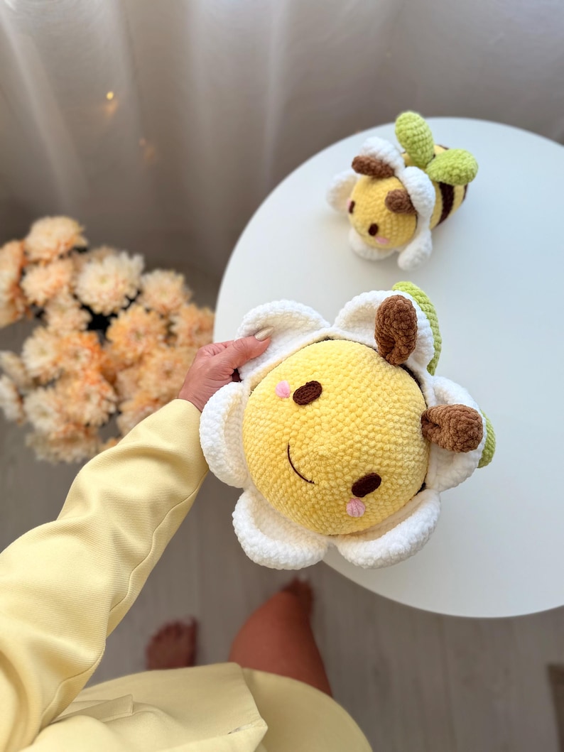 K&ouml;nnte beinhalten: Zwei handgefertigte H&auml;keltiere: eine gelbe Biene und eine gelbe Blume mit einem l&auml;chelnden Gesicht. Die Blume hat wei&szlig;e Bl&uuml;tenbl&auml;tter und braune Akzente. Die Biene hat braune und gr&uuml;ne Details. Beide liegen auf einer wei&szlig;en Oberfl&auml;che.