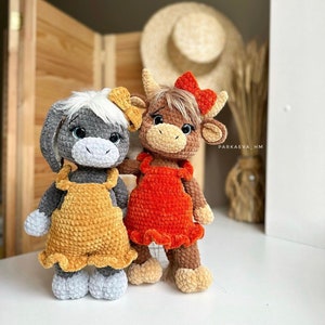 May include: Two crocheted stuffed animals, one gray with yellow overalls and the other brown with red overalls. Both animals have large eyes and are standing on a white surface.