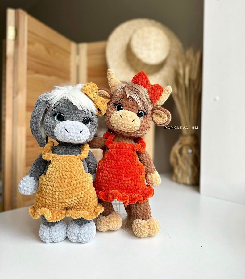May include: Two crocheted stuffed animals, one gray with yellow overalls and the other brown with red overalls. Both animals have large eyes and are standing on a white surface.