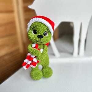 Peut inclure: Une poup&eacute;e en peluche crochet&eacute;e verte avec de grands yeux marron et un nez cousu noir. La poup&eacute;e porte un bonnet de P&egrave;re No&euml;l rouge et blanc et tient une canne en sucre ray&eacute;e rouge et blanche dans ses bras. La texture du fil est douce et duveteuse.
