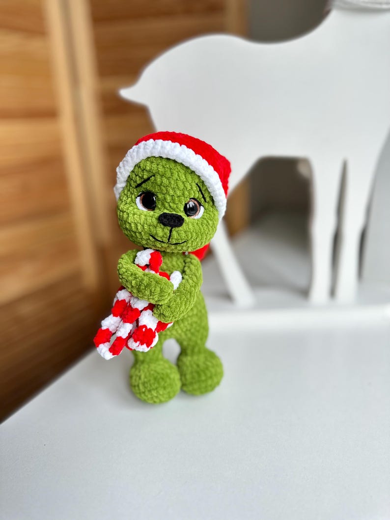 Peut inclure: Une poup&eacute;e en peluche crochet&eacute;e verte avec de grands yeux marron et un nez cousu noir. La poup&eacute;e porte un bonnet de P&egrave;re No&euml;l rouge et blanc et tient une canne en sucre ray&eacute;e rouge et blanche dans ses bras. La texture du fil est douce et duveteuse.