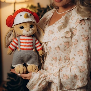 Peut inclure: Une poupée lapin au crochet faite à la main, vêtue d'une chemise rayée rouge et blanche, d'un short gris et d'un chapeau de bonhomme de neige. Le lapin a de grands yeux expressifs et tient une carotte. C'est un jouet doux et en peluche.
