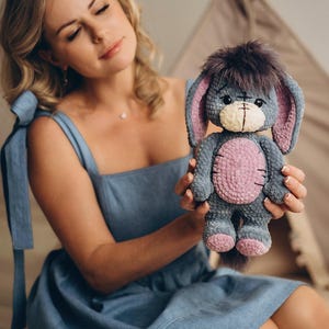 Pu&ograve; includere: Una donna con un vestito blu tiene in mano un peluche all'uncinetto che assomiglia a un asino. L'asino &egrave; grigio con dettagli rosa e ha una coda soffice.