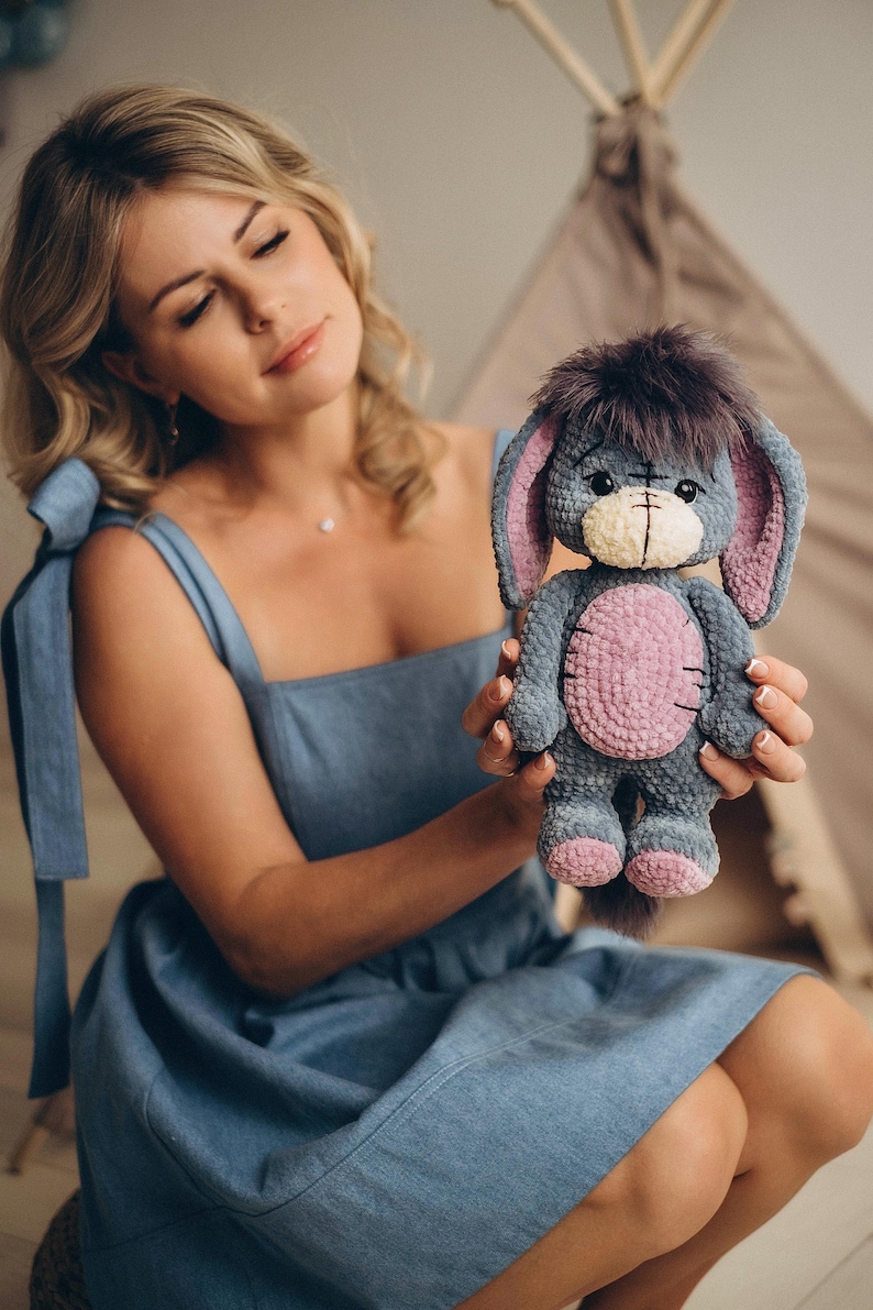 Pu&ograve; includere: Una donna con un vestito blu tiene in mano un peluche all'uncinetto che assomiglia a un asino. L'asino &egrave; grigio con dettagli rosa e ha una coda soffice.