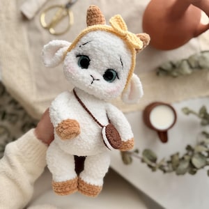 May include: A white crocheted plush lamb toy with brown hooves and ears. The lamb wears a yellow headband with a small bow, has large blue eyes with long eyelashes, and carries a small brown and white round cookie-shaped bag on a thin strap.