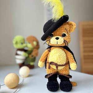 Pode incluir: Um gato de pelúcia de crochê vestindo um chapéu preto com uma pena amarela, uma capa marrom e botas pretas. O gato é marrom e amarelo com olhos pretos e um nariz preto.