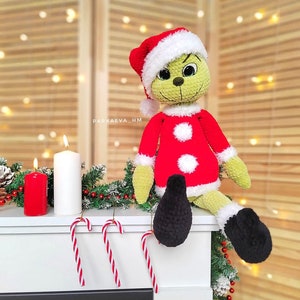 May include: A handmade green Grinch doll wearing a red Santa suit with white trim and a matching hat. The doll has black shoes and is sitting on a white surface with candy canes and candles.