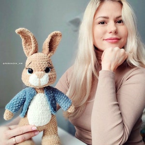 Puede incluir: Un conejito de crochet marr&oacute;n que lleva una chaqueta vaquera azul. El conejito tiene pelo blanco en el pecho y el vientre. El conejito est&aacute; de pie sobre una mesa frente a una mujer con el pelo largo y rubio.