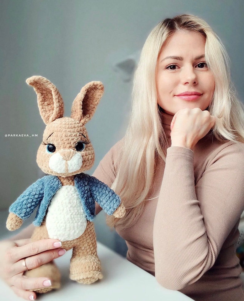 Puede incluir: Un conejito de crochet marr&oacute;n que lleva una chaqueta vaquera azul. El conejito tiene pelo blanco en el pecho y el vientre. El conejito est&aacute; de pie sobre una mesa frente a una mujer con el pelo largo y rubio.