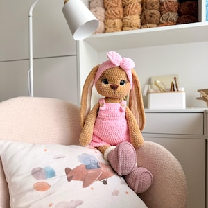 May include: A handmade crochet bunny doll with long ears, wearing a pink headband, a pink overall outfit, and mauve boots. The plush toy has dark eyes and a brown nose, sitting on a textured chair with a pillow featuring an airplane illustration.