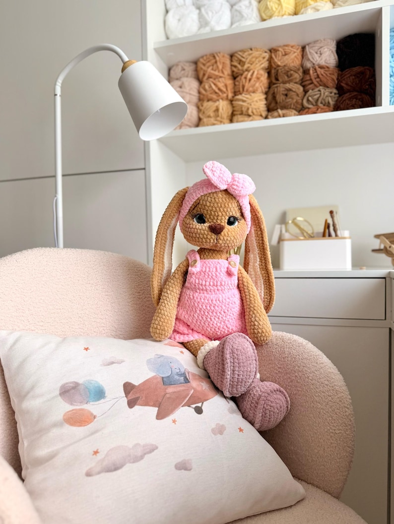 May include: A handmade crochet bunny doll with long ears, wearing a pink headband, a pink overall outfit, and mauve boots. The plush toy has dark eyes and a brown nose, sitting on a textured chair with a pillow featuring an airplane illustration.