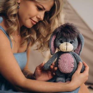 Pu&ograve; includere: Una donna con un vestito blu tiene in mano un peluche grigio lavorato a uncinetto con dettagli rosa. L'animale ha grandi orecchie e una testa pelosa.