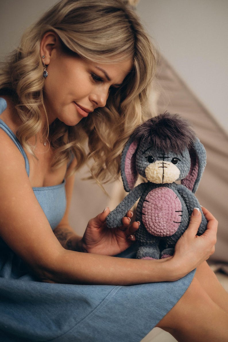 Pu&ograve; includere: Una donna con un vestito blu tiene in mano un peluche grigio lavorato a uncinetto con dettagli rosa. L'animale ha grandi orecchie e una testa pelosa.