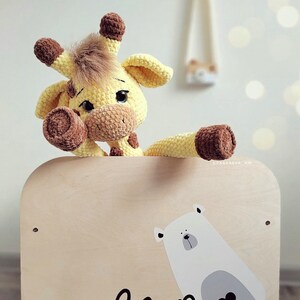 May include: A crocheted yellow giraffe toy with brown accents is peeking over the top of a wooden toy box. The box has a white polar bear illustration with a heart on its chest and the word "bear" written below it.