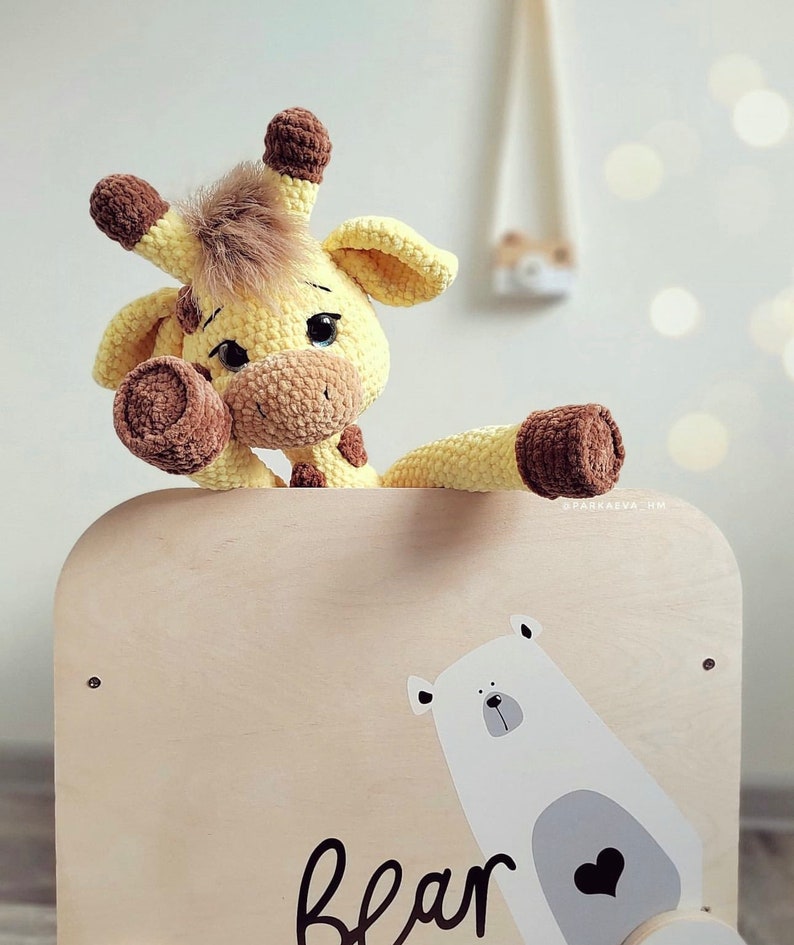 May include: A crocheted yellow giraffe toy with brown accents is peeking over the top of a wooden toy box. The box has a white polar bear illustration with a heart on its chest and the word "bear" written below it.
