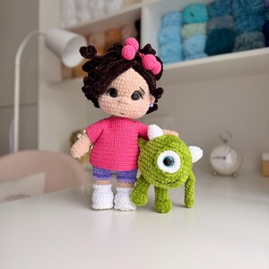 May include: A handmade crochet doll with dark brown curly hair adorned with two pink bows, wearing a bright pink t-shirt, purple shorts, and white shoes. Beside the doll stands a small, round, green one-eyed monster crochet plush toy.